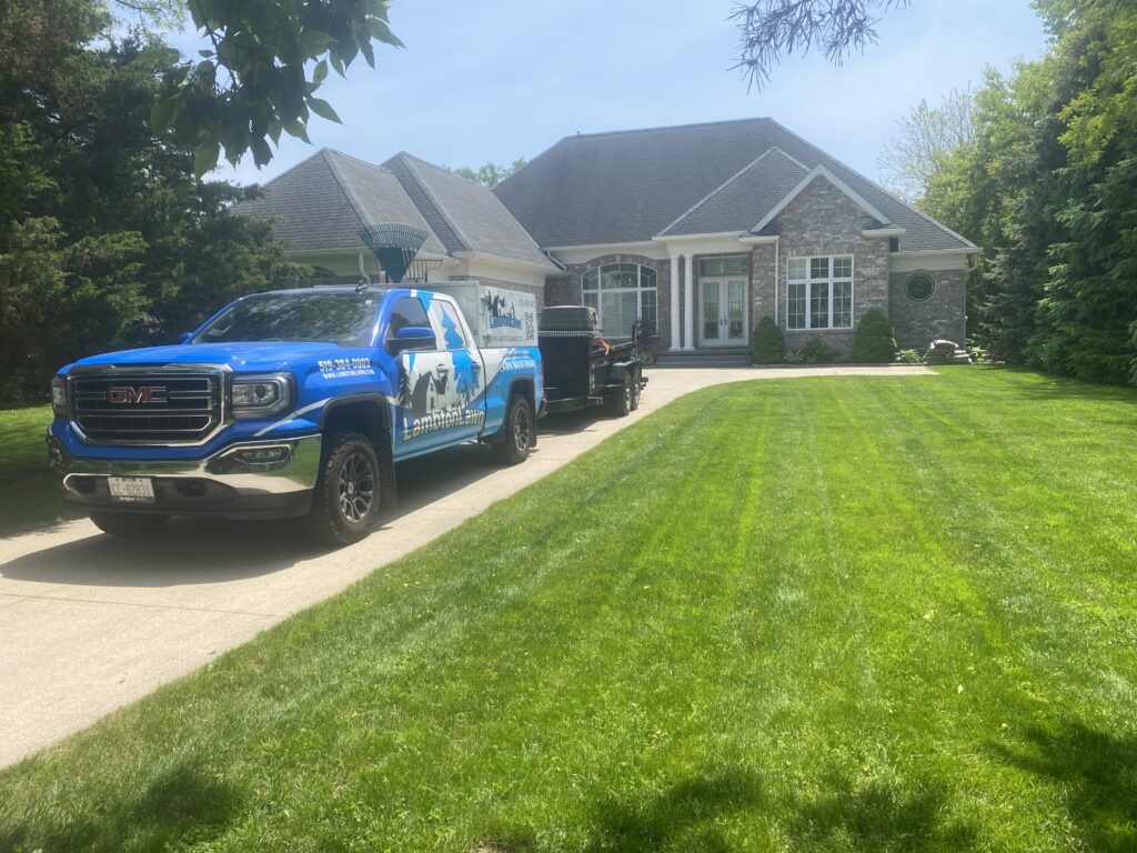 Brightly lit image of a well-maintained residential lawn featuring a blue GMC truck with landscaping branding parked on a driveway. The house in the background showcases elegant architecture and manicured greenery, highlighting professional landscaping services.
