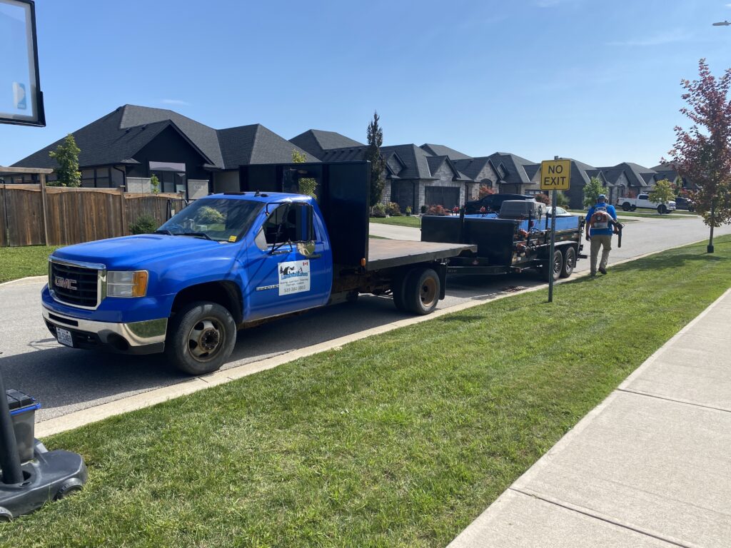 Blue landscaping truck parked on a residential street, with a worker preparing equipment for yard maintenance, showcasing professional landscaping services in a suburban area.