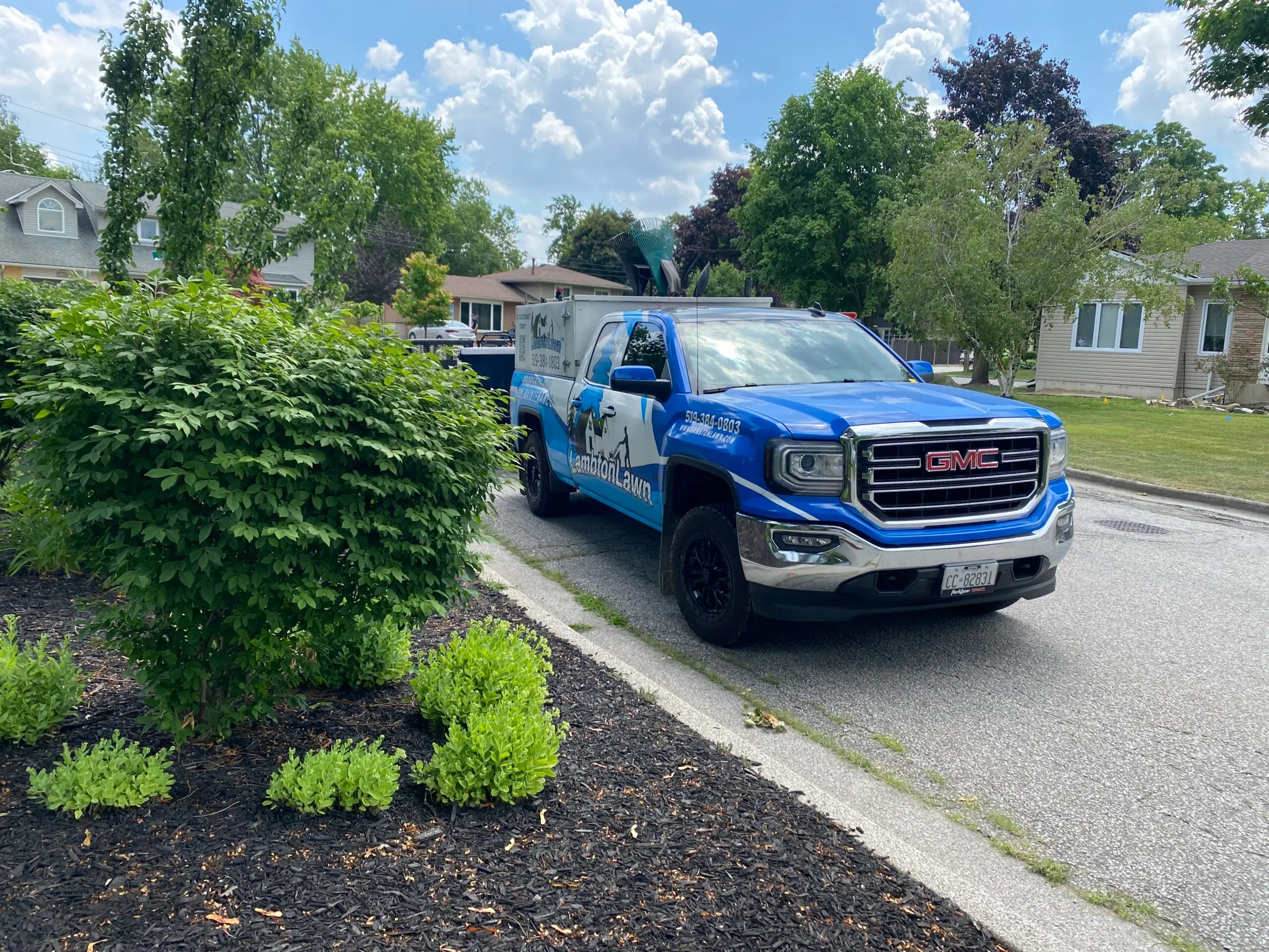 Landscaping service vehicle parked on a residential street, showcasing vibrant green shrubs and well-maintained flower beds with black mulch, under a clear blue sky.